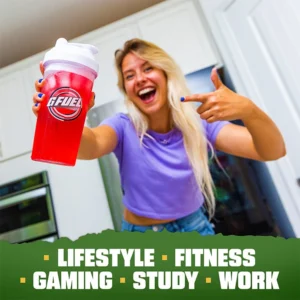 Smiling woman in a purple shirt holds up a red GFUEL shaker in a kitchen, gesturing to it with excitement.